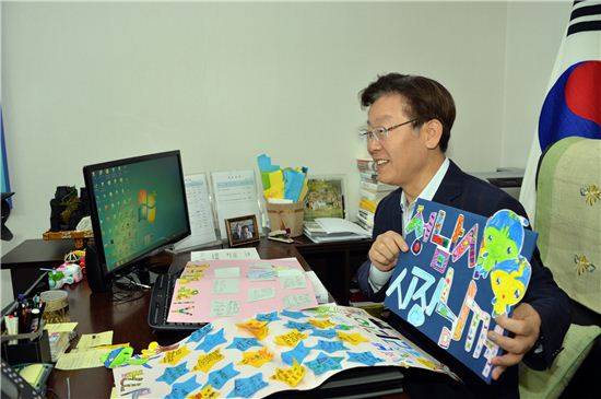 이재명 성남시장이 집무실에 앉아서 단식과 입원 기간동안 보내준 시민들과 어린이들의 격려 문구를 들어보이며 환하게 웃고 있다.