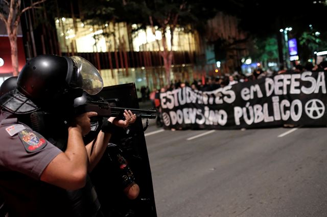 브라질 상파울루에서 23일(현지시간) 자이르 보우소나루 정부의 교육정책을 반대하는 시위가 열리고 있는 가운데 시위대 해산을 위해 배치된 진압경찰들이 '공교육을 옹호하는 학생들'이라고 적힌 현수막을 들고 행진 중인 시위대 앞에 서 있다.  로이터 연합뉴스