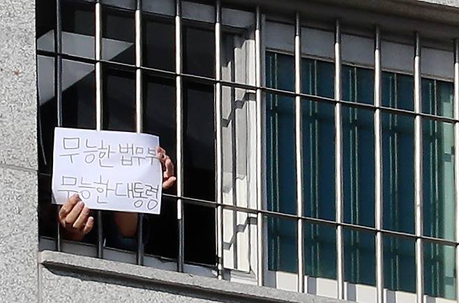 6일 오후 서울 송파구 동부구치소에서 한 수용자가 '무능한 법무부 무능한 대통령'이라고 쓴 종이를 창문 밖 취재진에게 내보이고 있다. 법무부는 이날 동부구치소 수용자 66명이 추가로 확진 판정을 받았다고 밝혔다. [연합]