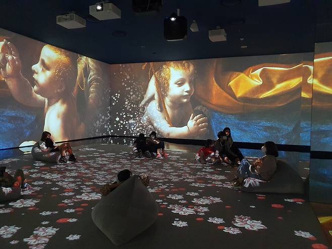 다빈치의 작품들이 사방 벽면에 투사된 몰입형 공간에서 관람객들이 빈백에 앉아서 작품을 즐기고 있다.