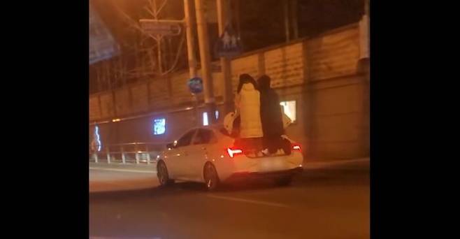 달리는 차량의 트렁크에 서서 한 손은 뚜껑을 붙잡고, 한 손은 서로의 팔짱을 낀 채 이동하는 모습.