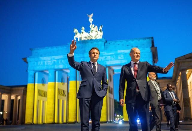 지난 9일 독일을 방문한 에마뉘엘 마크롱(왼쪽) 프랑스 대통령이 올라프 숄츠 독일 총리와 함께 베를린 브란덴부르크문 앞에서 시민들에게 인사를 하고 있다. 브란덴부르크문은 유럽의 날을 맞아 우크라이나 국기 색상 조명으로 장식됐다. 베를린=AP 뉴시스