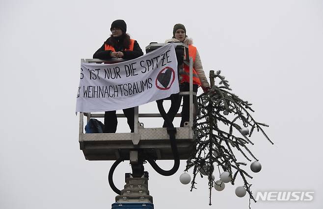 [베를린=AP/뉴시스]독일의 기후운동단체 '마지막 세대'의 운동가 2명이 21일 베를린의 상징 브란덴브루크문 옆 파리 광장에 세워진 15m 높이의 크리스마스 트리 꼭대기 약 2m 부분을 톱으로 잘라낸 후 "이는 단지 크리스마스 트리의 꼭대기일 뿐"이라고 쓴 현수막을 들고 있다. 2022.12.21