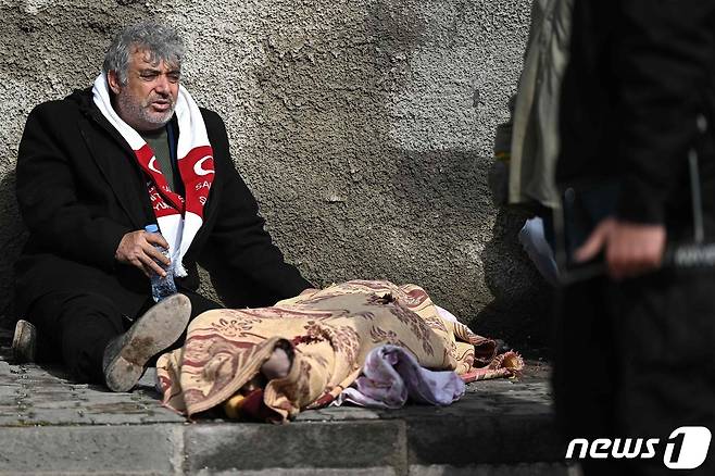 튀르키예 카라만마라슈의 건물 잔해 옆에서 한 남성이 담요를 씌운 아이 시신 옆에 앉아 있다. ⓒ AFP=뉴스1