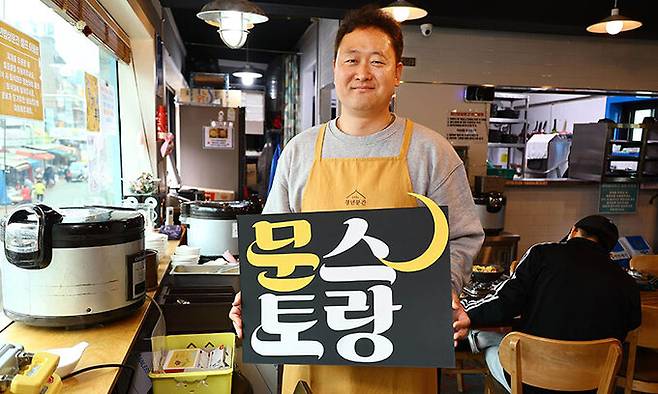 이문수 신부가 지난 4일 ’청년밥상 문간’ 식당에서 앞치마를 두른 채 ‘문스토랑(문간+레스토랑)’ 팻말을 들고 포즈를 취했다. 지금은 주야간 홀 담당 아르바이트생이 한 명씩 있어 이 신부는 식당보다 주로 옆 북카페에서 업무를 본다. 남제현 선임기자