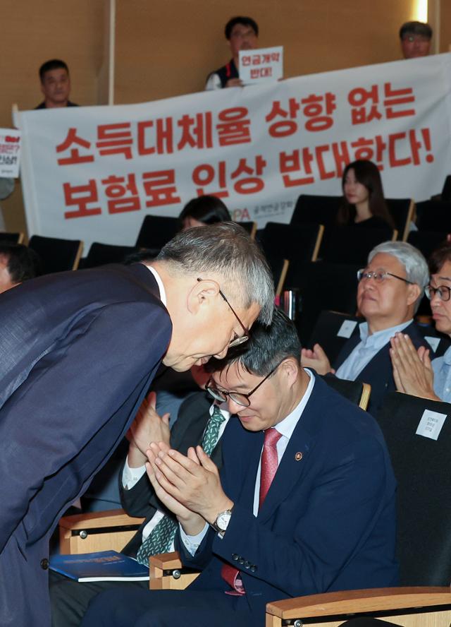 김용하 재정계산위원장이 1일 서울 코엑스에서 열린 국민연금 재정계산위원회 공청회에서 참석자들에게 인사하고 있다. 김용하 위원장 너머로 노동·시민·사회단체들로 구성된 공적연금강화국민운동(연금행동) 관계자들이 규탄 피켓을 들고 있다. 연합뉴스