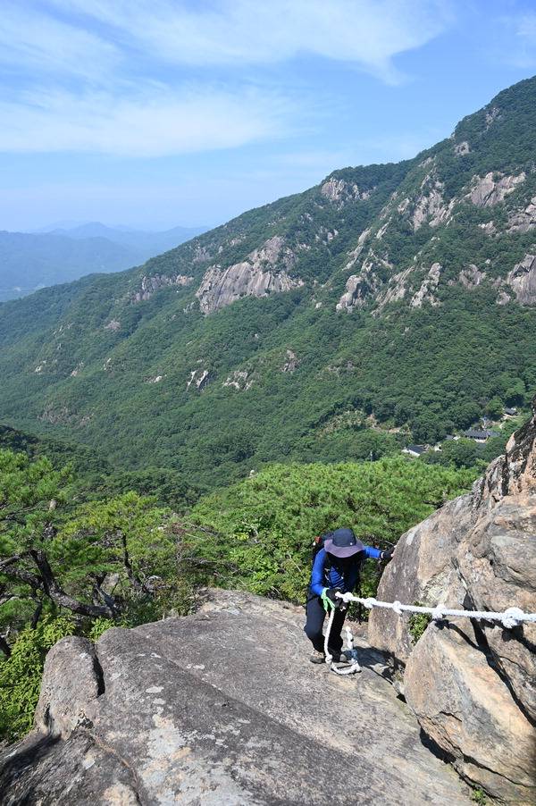 남근석을 지나 로프가 묶인 암릉을 오르는  취재팀 뒤로 배바위와 무암사가 보인다.