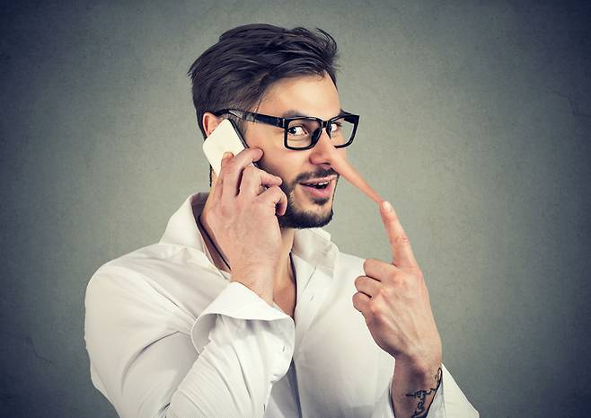 Young handsome man with long nose of liar cheating while speaking on smartphone and looking at camera.