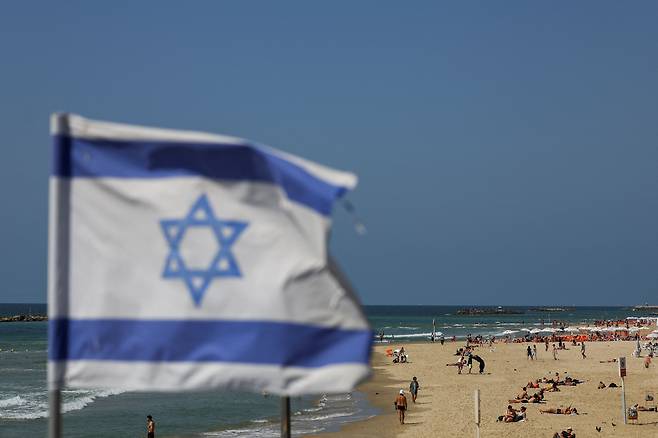 이란의 공습이 끝난 14일(현지시각) 오전 이스라엘 텔아비브 해변에서 이스라엘 국기 뒤로 사람들이 쉬고 있다. /로이터·연합뉴스