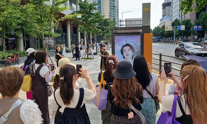 서울 용산구 하이브 사옥 앞 버스정류소에서 BTS 팬들이 데뷔 11주년을 축하하는 광고 앞에서 기념 사진을 찍고 있다.