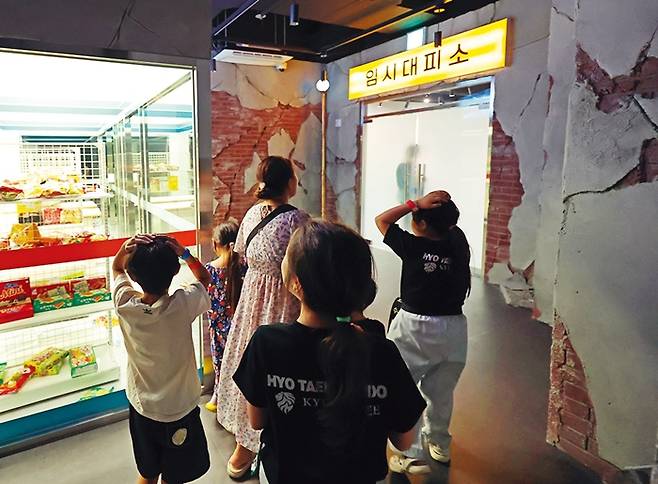 참가자들이 지진으로 금이 가고 무너진 건물 사이를 지나 임시대피소로 대피하고 있다. 대피하는 도중에도 여진으로 마트 내부가 흔들리는 모습을 볼 수 있다.