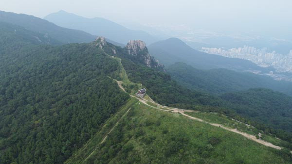 금정산성 4망루에서 드론을 날려서 찍어 본 전망이다. 사진 가운데 누각이 4망루이며 아래로 중성이 내려가는데 근교산 취재팀 하산길이다. 망루 뒤로 의상봉과 오른쪽 암봉은 부산 산악인의 암벽 등반 코스로 인기를 끄는 무명암이며, 멀리 계명봉과 금정구 청룡동이 보인다.