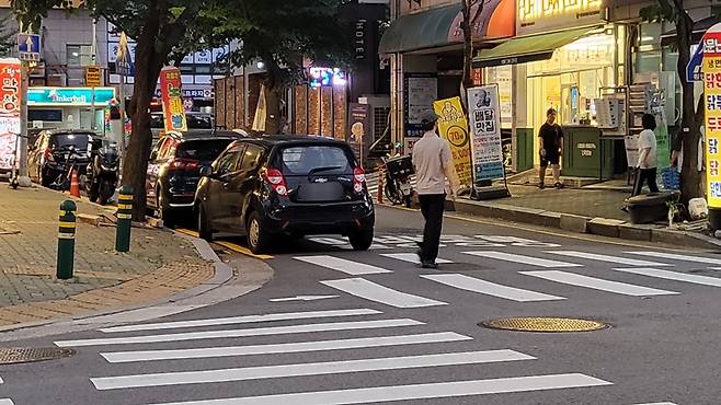 교차로 모퉁이, 횡단보도에 걸쳐서 주차한 차량과 횡단보도를 건너는 행인. 신고할 때는 배경도 잘 나오게 찍어서 신고해야 한다.