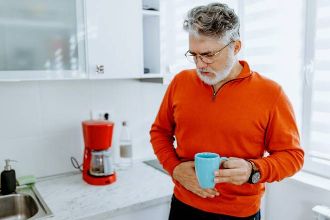 Worried mature Caucasian man standing in kitchen with hand on his painful stomach while holding coffee