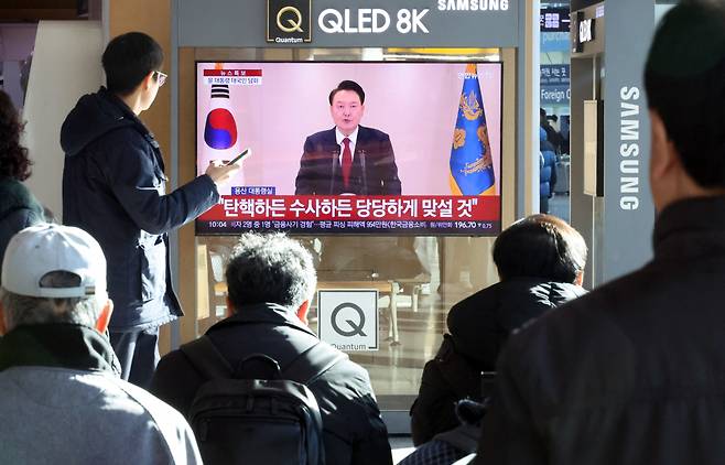 Citizens watch President Yoon Suk Yeol deliver a public address on television at Seoul Station on Thursday morning. (Yonhap)