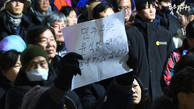 3일 계엄 직후, 국회 앞으로 모인 시민들은 무장한 계엄군을 맨몸으로 막아냈다. 한 시민이 '민주주의는 윤석열 따위에게 굴복하지 않는다'라고 적은 종이를 들고 있는 모습.