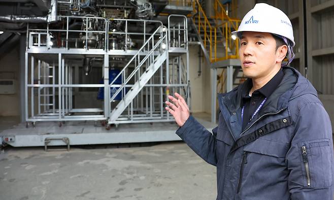 전준수 한국항공우주연구원 선임연구원이 나로우주센터 엔진 시험동에서 진행 중인 엔진 수락 연소시험에 대해 설명하고 있다. 사진 속 스탠드에는 누리호 5호기의 첫 번째 1단 엔진이 실험을 준비하고 있다./한국항공우주연구원