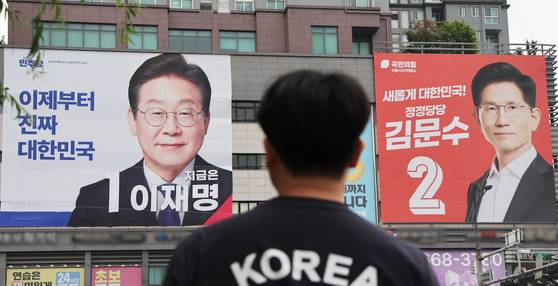 A man wearing a ″Korea″ shirt stands in front of posters for Lee Jae-myung, left, and Kim Moon-soo