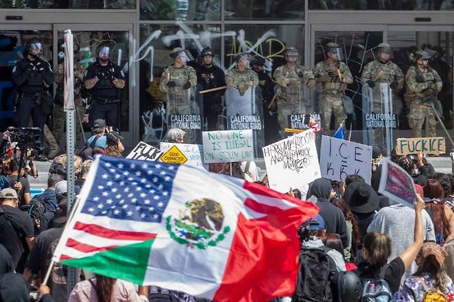 2025년 6월 9일, 로스앤젤레스에서 열린 시위에서 시위대가 팻말과 미국-멕시코 혼합 국기를 들고 연방청사 앞을 지키는 캘리포니아 주방위군 병력과 대치하고 있다. (사진=AFP)