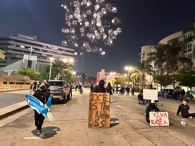 9일 밤 미국 캘리포니아주 로스앤젤레스에서 트럼프 행정부의 이민자 추방 정책에 항의하는 시위대가 미 이민 단속국은 '새로운 나치 친위대(SS)'에 빗대는 팻말 등을 들고 경찰과 대치하고 있다. 밤하늘에서 시위대가 쏘아올린 폭죽이 터지고 있다./오로라 특파원