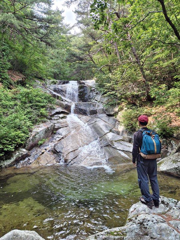 경남 함양 ‘오르GO 함양’ 산악 완등 인증 네 번째 월봉산은 큰목재골로 올라 새명골로 하산하는 계곡 산행이다. 새명골의 다박골(따박골) 폭포에 내려선 취재팀이 높이 20m에서 떨어지는 폭포의 물줄기를 보며 무더위를 식히고 있다.