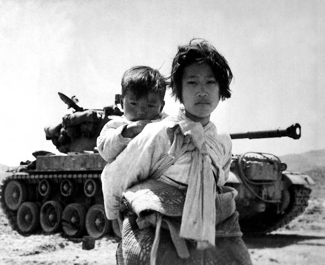 With her brother on her back, a war-weary Korean girl tiredly trudges by a stalled M-26 tank, at Haengju, Korea, on June 9, 1951. [U.S. NATIONAL ARCHIVES]
