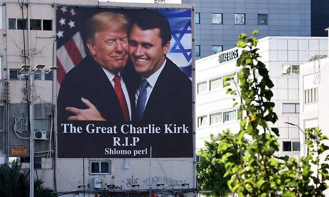 13일(현지시각) 이스라엘 텔아비브의 한 건물 바깥벽에 지난 10일 피살된 미국 청년 우파 논객 찰리 커크를 추모하는 사진이 걸렸다. 사진 속 커크(오른쪽)는 도널드 트럼프 미국 대통령과 볼을 맞대고 껴안고 있다. 텔아비브/AFP 연합뉴스