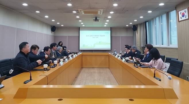 회의실에서 여러 사람들이 테이블에 앉아 있고, 스크린에는 흐릿한 텍스트가 표시된 사진이다.테이블 위에는 컵이 놓여 있고, 회의 참가자들은 발표를 듣고 있는 듯한 모습이다.