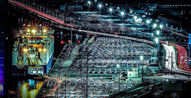 울산 현대차 수출부두의 야경은 끝없이 늘어선 차량들과 불 밝힌 선적선이 어우러져, 다시 달아오를 자동차 수출을 예고하는 듯한 풍경을 보여준다.  울산 현대자동차 수출부두에는 막 생산을 마친 차량들이 촘촘히 주차돼 거대한 흰 물결을 이루고, 부두 끝에서는 거대한 자동차 운반선이 선적을 기다리며 불을 밝힌다.  고가도로와 항만을 따라 이어지는 차량의 테일램프와 가로등 불빛이 붉고 흰 실선처럼 겹쳐지며, 세계 각지로 향하는 '수출 행렬'의 활기를 시각적으로 드러낸다.  수출 증가 기대감국내 완성차 업계는 내년 생산과 수출이 모두 소폭 증가할 것으로 내다보며, 특히 친환경차 라인업 확대로 글로벌 판매 회복을 기대하고 있다.  울산항 역시 자동차와 철강 화물을 중심으로 물동량이 개선되는 흐름을 보이고 있어, 부두를 가득 메운 신차 행렬이 머지않아 실제 수출 증가로 이어질 것이라는 전망에 힘을 싣는다. 김재훈 기자