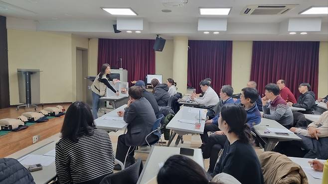 강당에서 한 여성이 심폐소생술 교육을 진행하고 있고, 사람들이 책상에 앉아 교육을 듣고 있는 사진이다. 책상 위에는 교육 자료로 보이는 종이들이 놓여 있다.