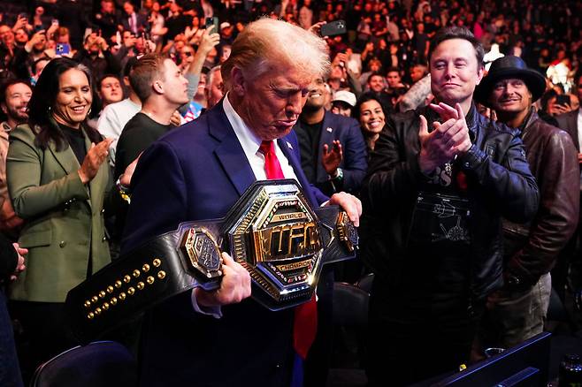 지난 2024년 11월 미국 뉴욕 메디슨스퀘어가든에서 열린 UFC 309 대회에서 헤비급 챔피언 존 존스의 벨트를 잠시 건네받고 감상중인 도널드 트럼프 미 대통령. 트럼프는 소문난 격투기, 프로레슬링 애호가다. 그 옆에는 당시 사이가 좋았던 일론 머스크 테슬라 CEO가 박수를 치고 있는 모습이다. 트럼프 미 행정부는 지난 해 12월 39개국에 대한 비자 제한 조치를 단행했지만, 올해 1월 15일 올림픽, 월드컵을 비롯해 UFC, WWE, MLB, NBA, MLS, F1 등 주요 프로대회의 출전선수와 코치진은 이 조치에서 제외하기로 했다. [게티이미지]