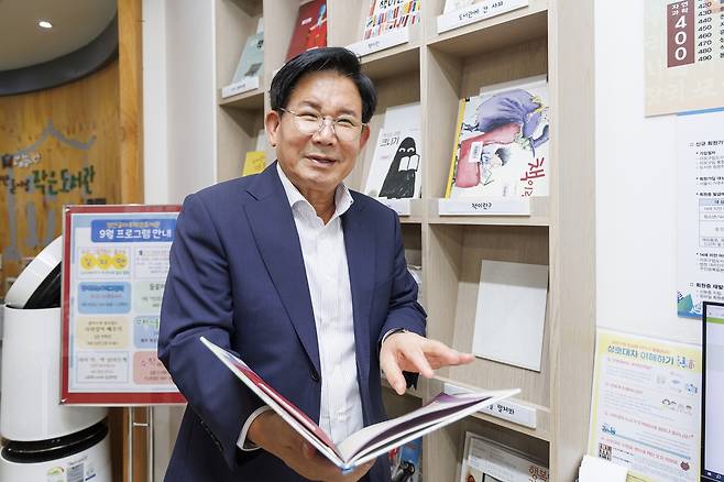 미소를 짓고 있는 박강수 마포구청장이 책을 들고 서 있는 사진이다. 배경에는 책들이 꽂혀있는 책장이 보인다.