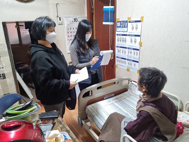 마스크를 착용한 두 명의 여성이 침대에 앉아있는 노인의 안부를 확인하고 있는 사진이다. 여성들은 각각 서류와 클립보드를 들고 있으며, 방 안에는 달력과 안내문이 보인다.