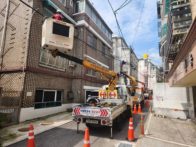전봇대와 건물 사이에 공중 케이블 정비 작업을 하는 차량이 위치한 사진이다. 작업자는 노란색 작업 차량의 높은 위치에 있는 상자 모양의 장비 안에서 케이블을 정비하고 있다.