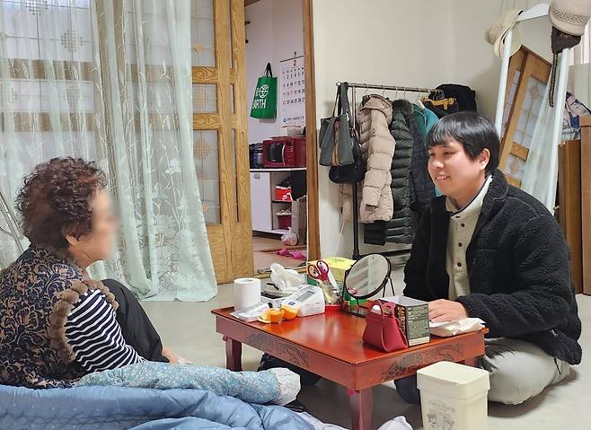 방 안에서 한 여성이 다른 여성과 마주 앉아 대화하고 있는 사진이다. 두 여성 사이에는 낮은 탁자가 놓여 있고, 탁자 위에는 귤과 거울 등이 놓여 있다.