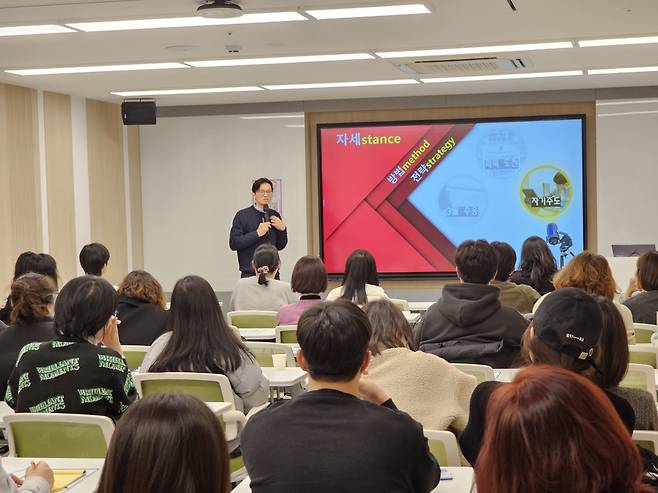 강사가 마이크를 들고 강의를 진행하고 있고, 학생들이 강의를 듣고 있는 사진이다. 화면에는 ‘자세 stance, 방법 method, 전략 strategy’라는 텍스트가 보인다.