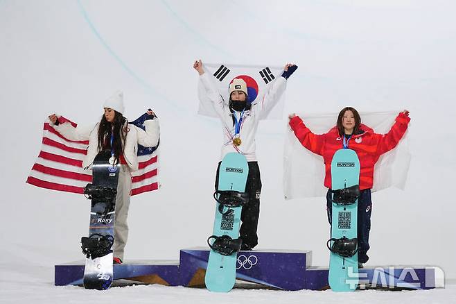 [리비뇨=AP/뉴시스] 최가온(가운데)이 12일(현지 시간) 이탈리아 리비뇨 스노파크에서 열린 2026 밀라노·코르티나담페초 동계올림픽 스노보드 여자 하프파이프 정상에 올라 태극기를 들고 포즈를 취하고 있다. 최가온은 1차 시기에서 크게 넘어져 충격을 안겼으나 3차 시기에서 90.25점을 획득해 강력한 우승 후보 클로이 김(미국, 왼쪽)을 제치고 금메달을 목에 걸었다. 최연소 금메달 기록도 갈아치운 그는 시상식 도중 애국가가 흘러나오자, 끝내 눈물을 흘렸다. 2026.02.13.