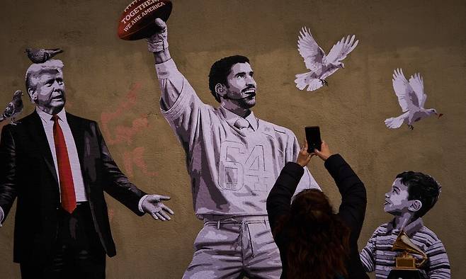 지난 17일(현지시각) 스페인 바르셀로나 도심 거리에서 한 여성이 배드 버니(가운데)의 슈퍼볼 공연 장면과 도널드 트럼프 미국 대통령(왼쪽)을 함께 그린 그래피티 벽화를 촬영하고 있다. AP 연합뉴스