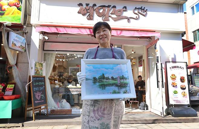 김은진씨가 2026년 3월 말로 영업을 마치는 자신의 빵집 앞에서 자신이 그린 어반스케치를 들어 보이고 있다. 뒤편 빵집 안에서 어반스케처들이 바깥 풍경을 스케치하고 있다.