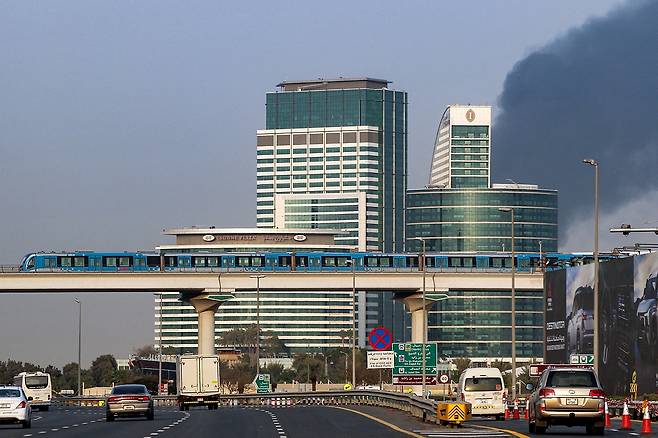 16일(현지시간) 두바이 국제공항 인근에서 발생한 화재로 인해 연기가 치솟는 가운데 두바이 지하철 열차가 고속도로 위의 차량들 위로 지나가고 있다. [AFP]