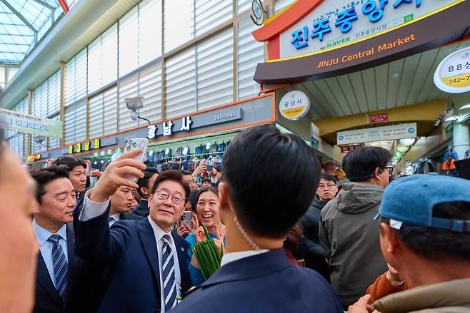 이재명 대통령이 25일 한국항공우주산업(KAI)에서 열린 KF-21 양산 1호기 출고식에 참석한 뒤 경남 진주시 진주중앙시장을 방문해 시민과 사진을 찍고 있다. 청와대 제공