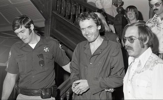 연쇄 범죄자 테드 번디가 1977년 미국 콜로라도 피트킨 카운티 법원에서 경찰의 호송을 받으며 이동하고 있다. 번디는 카메라를 피하지 않고 늘 웃음을 지었으며, 당시 재판을 기다리던 중 두 차례 탈출해 사회에 큰 충격을 안겼다. AP 연합뉴스 자료사진