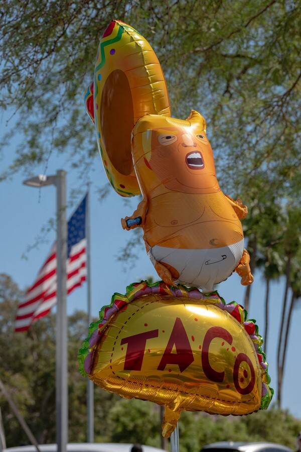 도널드 트럼프 미국 대통령이 지난 17일(현지시간) 애리조나주 피닉스에서 열린 보수 단체 '터닝 포인트 USA'의 행사 '붉은 담을 쌓자' 참석차 방문하는 것을 반대하는 사람이 드림 시티 교회 바깥에서 트럼프를 아기로 묘사하고 '트럼프는 항상 겁을 먹고 물러선다'(TACO)고 적힌 타코 볼 모양의 풍선을 들고 있다. 터닝 포인트 USA는 지난해 9월 암살된 찰리 커크가 창립해 이끌던 단체다. 2026.4.17. 피닉스 AFP 연합뉴스