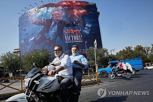 2026년 8월 24일 이란 테헤란에서 오토바이 운전자들이 '위대한 승리! 호르무즈 해협'이라는 문구와 함께 도널드 트럼프 미국 대통령이 폭발 속에서 물에 잠긴 모습이 그려진 대형 광고판 앞을 지나고 있다. [AP 연합뉴스. 재판매 및 DB 금지]