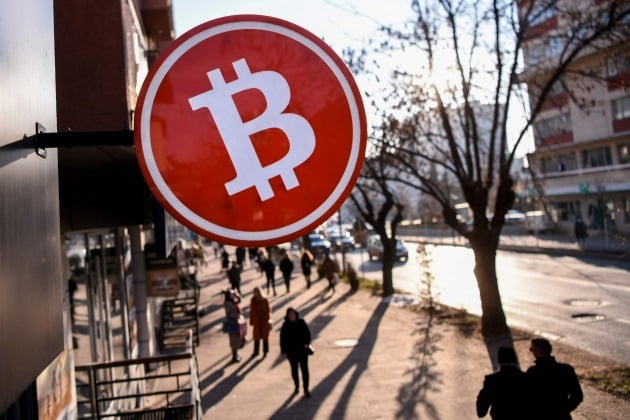 Pedestrians walk under a Bitcoin cryptocurrency symbol displayed at a cryptocurrency exchange shop in Pristina on January 17, 2022. AFP연합뉴스