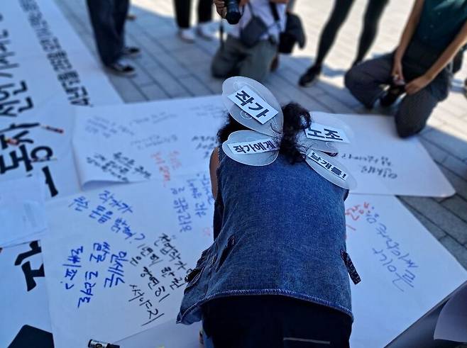 ▲2024년 6월26일 서울국제도서전이 열린 서울 강남구 코엑스 앞, 작가들이 노동권에 날개를 달자는 취지의 날개 모형을 부착하고 퍼포먼스를 벌이고 있다. 출판사로부터 들어온 부당한 발언을 종이에 적은 뒤 찢어버리기도 했다. 사진=작가노조준비위