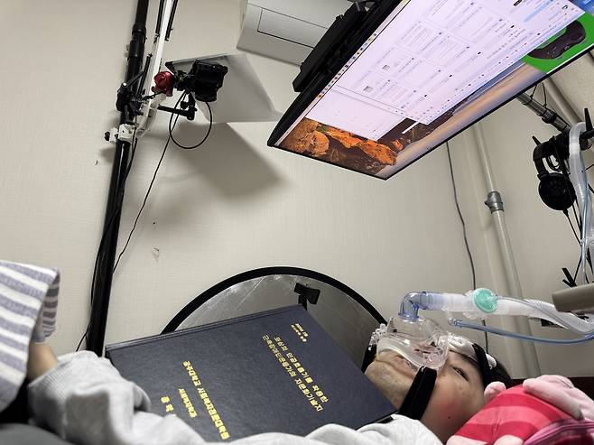 Jang Ik-seon holds his completed master’s thesis bound in print — written through nearly half a million blinks — lying beneath the eye-tracking system that made it possible at his home in Gwangju. (Moon Joon-hyun/The Korea Herald)