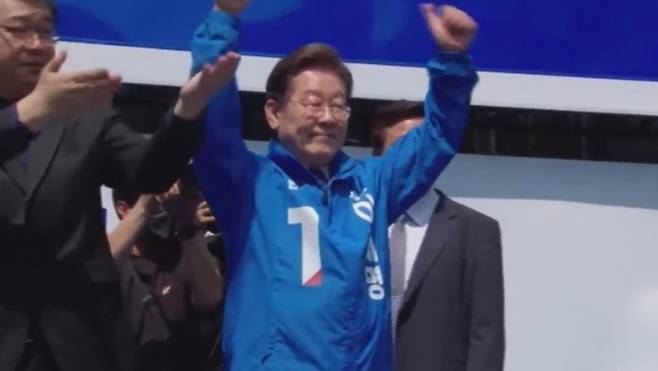 출정식 무대에 오른 이재명 더불어민주당 대선 후보가 파란 점퍼를 입고 두 팔을 들어 보이며 지지자들의 환호에 응답하고 있다. 유세 현장에는 안전 확보를 위한 방호 조치가 대폭 강화됐다. ('델리민주' 캡처)