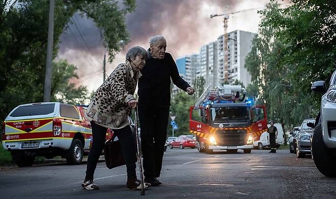 4일(현지시각) 러시아가 우크라이나 키이우에 드론과 미사일 공세를 가하면서, 아파트 뒤로 연기가 피어오르는 가운데 노부부가 길을 건너고 있다. UPI연합뉴스