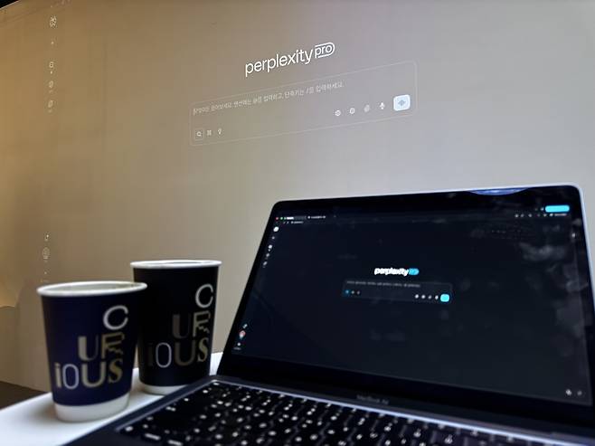 A laptop running Perplexity Pro sits in front of a wall-sized projection of the same interface, alongside coffee cups branded with the cafe’s name, Curious. (Moon Joon-hyun/The Korea Herald)